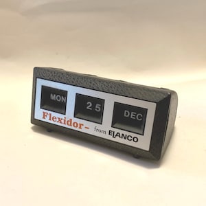 May include: A black desk calendar with white and silver accents. The calendar displays the date "25 DEC" and the day of the week "MON". The calendar is labelled "Flexidor - from ELANCO".