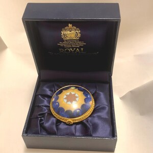 May include: A round, blue and gold trinket box with a white star and gold rays. The box is in a blue velvet box with the words "By Appointment to Her Majesty Queen Elizabeth II Manufacturers of China and Porcelain The Worcester Royal Porcelain Company Worcester ROYAL" on the inside of the lid.