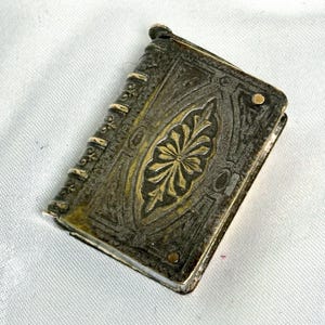 May include: A small, rectangular metal book-shaped object with a dark, tarnished finish and gold-toned accents. The cover features an embossed central floral motif, while the spine displays raised bands with intricate decorative detailing.