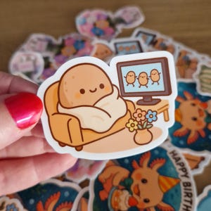 May include: A sticker featuring a cartoon potato character relaxing on a couch, watching TV. The potato is wrapped in a blanket. A small vase of flowers sits on a table next to the TV. Other stickers are in the background.