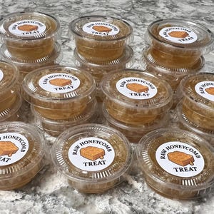 May include: A collection of small, clear plastic containers filled with raw honeycomb. Each container has a white label with the text "RAW HONEYCOMB TREAT" and a graphic of a honeycomb. The containers are stacked on top of each other.