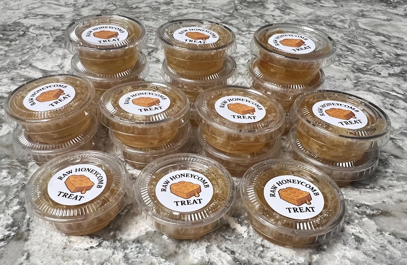 May include: A collection of small, clear plastic containers filled with raw honeycomb. Each container has a white label with the text "RAW HONEYCOMB TREAT" and a graphic of a honeycomb. The containers are stacked on top of each other.