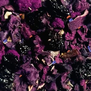 May include: Close-up of a mix of dried tea leaves and flowers. The image features a blend of deep purple, black, and brown elements, with small blue and white accents. The texture appears to be a mix of dried petals and berries.