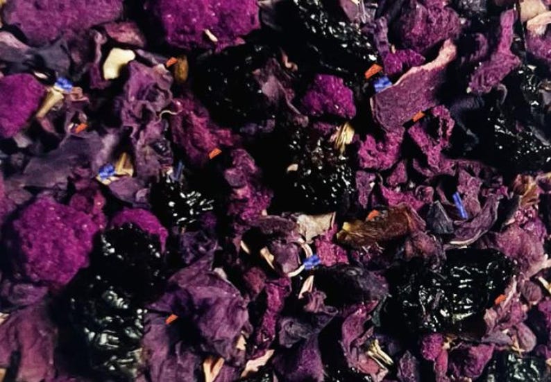 May include: Close-up of a mix of dried tea leaves and flowers. The image features a blend of deep purple, black, and brown elements, with small blue and white accents. The texture appears to be a mix of dried petals and berries.