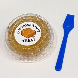 May include: A clear plastic container filled with raw honeycomb, labeled "RAW HONEYCOMB TREAT." A blue plastic spoon rests beside the container. The honeycomb is a golden yellow color.