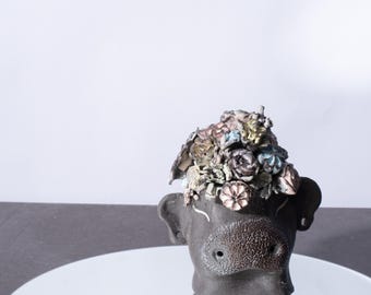 Puede incluir: Una escultura de cerámica negra de una cabeza con una corona de flores de colores. La escultura está sentada sobre un espejo redondo de plata.