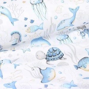 May include: White fabric with a repeating pattern of aquatic life. The design features blue whales, narwhals, fish, jellyfish, and sea turtles. The illustrations are in a watercolor style with gray and gold accents.