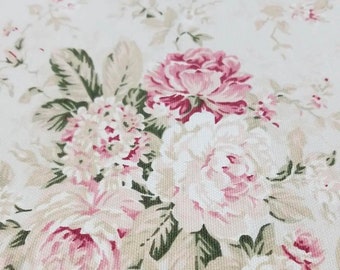 May include: A fabric featuring a floral pattern with large pink and white roses, smaller clusters of white blossoms, and muted green leaves set against a cream-colored background. The textile displays a visible woven texture throughout the surface.