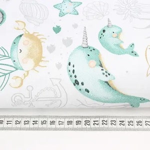 May include: White fabric with a repeating pattern of sea creatures, including narwhals, crabs, starfish, and fish. The narwhals are light blue with black spots and a horn. The crab is tan and light blue. A ruler is at the bottom.
