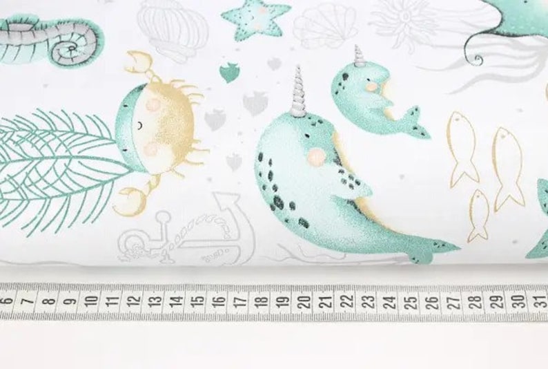May include: White fabric with a repeating pattern of sea creatures, including narwhals, crabs, starfish, and fish. The narwhals are light blue with black spots and a horn. The crab is tan and light blue. A ruler is at the bottom.