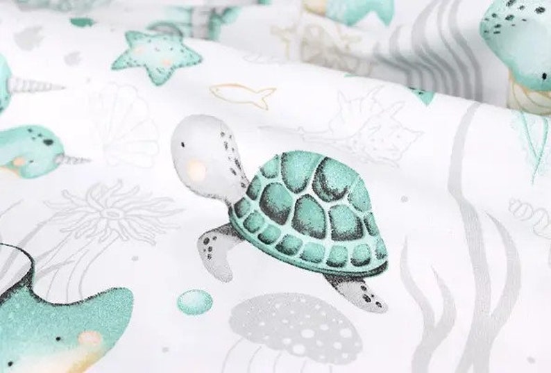 May include: A white fabric with a repeating pattern of sea creatures, including a turtle, starfish, and narwhal. The turtle is the focal point, with a teal shell and light gray body. Other sea life illustrations are in teal and gray.