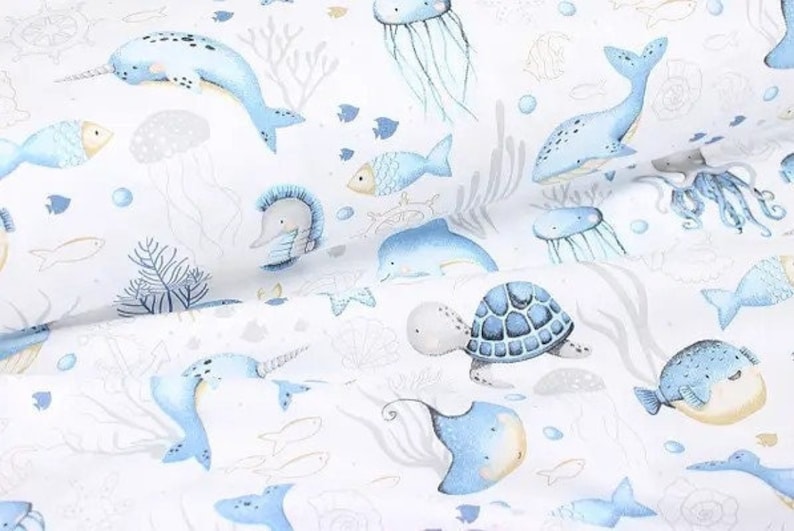 May include: White fabric with a repeating pattern of aquatic life. The design features blue whales, narwhals, fish, jellyfish, and sea turtles. The illustrations are in a watercolor style with gray and gold accents.