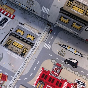 May include: A gray and white fabric with a repeating pattern of a city street scene. The fabric features buildings, cars, trees, and a fire station. The text "FIRE STATION" is printed on the fire station building.