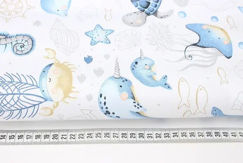 May include: White fabric with a repeating pattern of sea creatures in blue and gold. The design includes narwhals, crabs, starfish, and fish. A ruler with inch measurements is along the bottom.