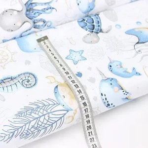 May include: White fabric with a repeating pattern of sea creatures in blue and beige. The design includes an octopus, seahorse, narwhal, and fish. A measuring tape shows the fabric's dimensions.