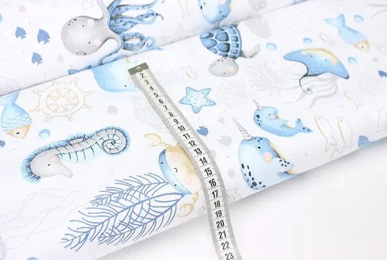 May include: White fabric with a repeating pattern of sea creatures in blue and beige. The design includes an octopus, seahorse, narwhal, and fish. A measuring tape shows the fabric's dimensions.