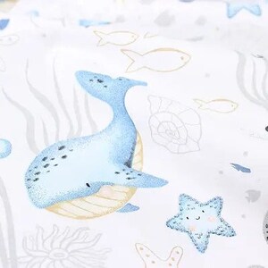 May include: White fabric with a repeating pattern of marine life. The design includes a light blue whale, starfish, and fish, along with sea plants and shells. The illustrations have a watercolor style.