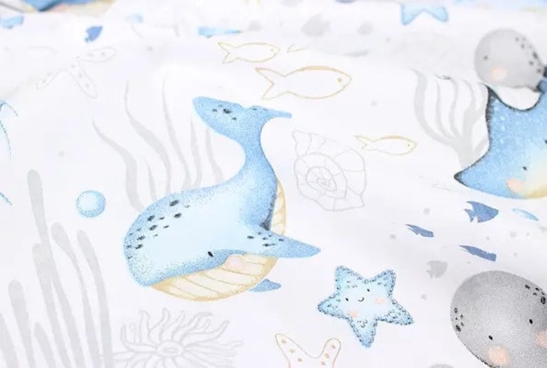 May include: White fabric with a repeating pattern of marine life. The design includes a light blue whale, starfish, and fish, along with sea plants and shells. The illustrations have a watercolor style.