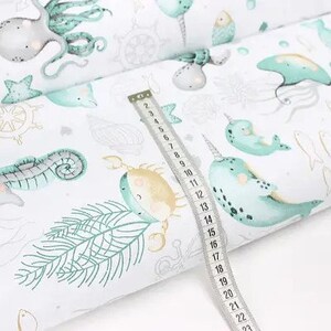 May include: White fabric with a sea life pattern, including octopuses, seahorses, narwhals, and fish in shades of teal and gray. A measuring tape shows a length of approximately 23 inches.