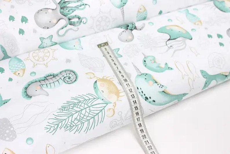 May include: White fabric with a sea life pattern, including octopuses, seahorses, narwhals, and fish in shades of teal and gray. A measuring tape shows a length of approximately 23 inches.
