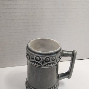 Puede incluir: Una pequeña taza de cerámica gris con un diseño decorativo alrededor de la parte superior e inferior. La taza tiene un asa.