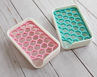 May include: Two white soap dishes with a honeycomb design. One dish is pink and the other is teal.