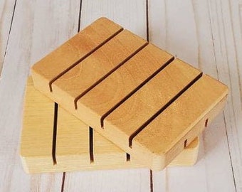 May include: Two wooden soap dishes. The top dish has five parallel slots, and the bottom dish has two slots. The wood has a light brown color. The background is a light wood grain.
