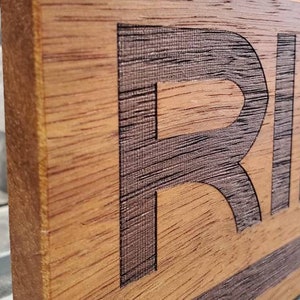 May include: A wooden sign with the word "RIDGID" engraved in black lettering. The sign has a dark brown stain and a black stripe across the bottom.