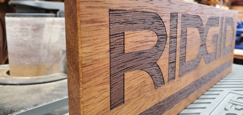 May include: A wooden sign with the word "RIDGID" engraved in black lettering. The sign has a dark brown stain and a black stripe across the bottom.