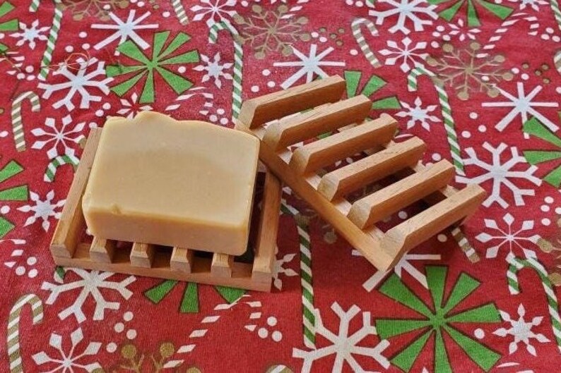 May include: A bar of soap in a wooden soap dish. The soap is a light beige color and the dish is made of natural wood. The soap dish is sitting on a red and white patterned fabric.