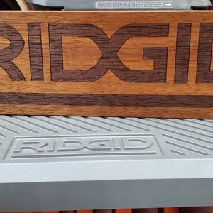May include: A wooden sign with the word "RIDGID" inlayed in a darker wood. The sign is sitting on a gray workbench with the word "RIDGID" printed on it.