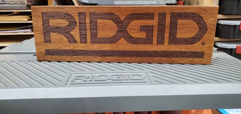 May include: A wooden sign with the word "RIDGID" inlayed in a darker wood. The sign is sitting on a gray workbench with the word "RIDGID" printed on it.