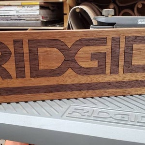 May include: A wooden sign with the word "RIDGID" engraved in a darker wood. The sign is made of two different types of wood, creating a contrasting effect.