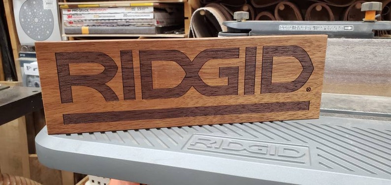 May include: A wooden sign with the word "RIDGID" engraved in a darker wood. The sign is made of two different types of wood, creating a contrasting effect.