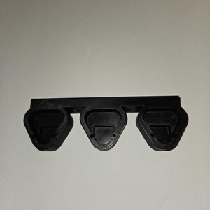 May include: A black plastic guitar pick holder with three triangular slots. The holder is mounted on a rectangular bar. The slots are designed to hold guitar picks, keeping them organized and accessible. The item is likely for musicians.