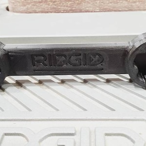 以下が含まれることがあります： 両端に星型の開口部のある黒いプラスチック製のレンチ。レンチには「RIDGID」のラベルが貼られています。