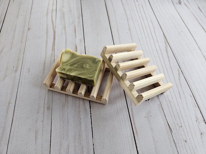 May include: Two wooden soap dishes, one with a green and white bar of soap on top. The dishes are made of natural wood and have a slatted design.