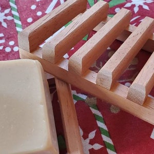 May include: A bar of natural soap resting on a wooden soap dish. The soap dish has a slatted design and is made of light brown wood. The soap is a light beige color and has a smooth, slightly textured surface.