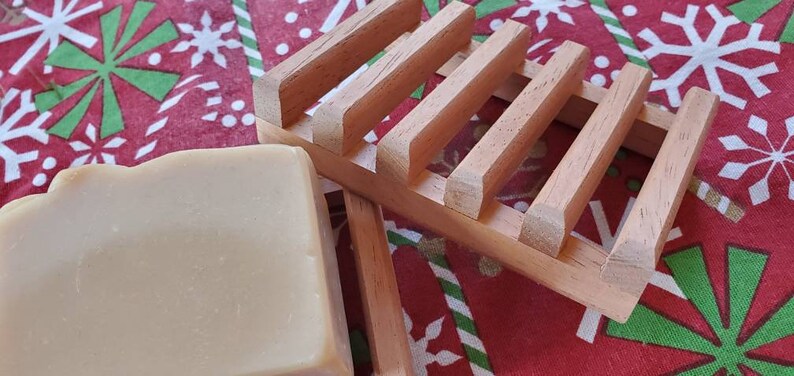 May include: A bar of natural soap resting on a wooden soap dish. The soap dish has a slatted design and is made of light brown wood. The soap is a light beige color and has a smooth, slightly textured surface.