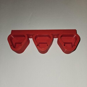 May include: A red, 3D-printed wall mount with three triangular-shaped holders. The holders are designed to accommodate small objects. The mount is made of a solid, opaque red plastic and is mounted on a white wall.