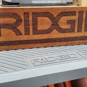 May include: A wooden sign with the word "RIDGID" engraved in dark brown letters. The sign is made of a light brown wood with a dark brown stripe running across the bottom.