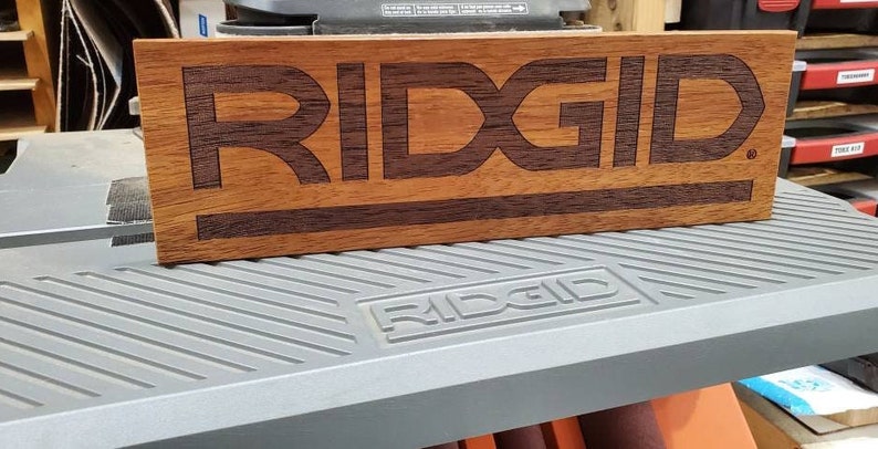 May include: A wooden sign with the word "RIDGID" engraved in dark brown letters. The sign is made of a light brown wood with a dark brown stripe running across the bottom.