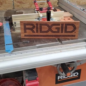 May include: A close-up of a wooden sign with the word "RIDGID" engraved in it. The sign is sitting on a table saw with a blue tape measure and a silver ruler.