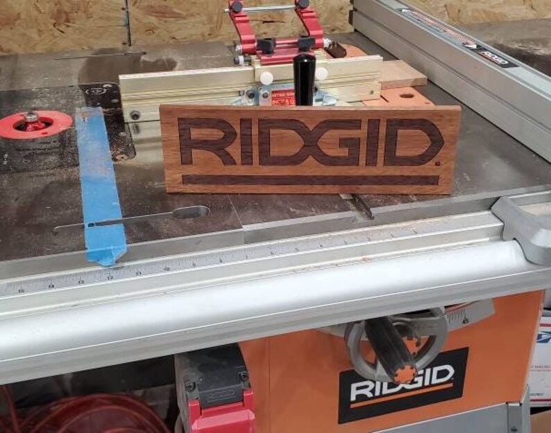 May include: A close-up of a wooden sign with the word "RIDGID" engraved in it. The sign is sitting on a table saw with a blue tape measure and a silver ruler.