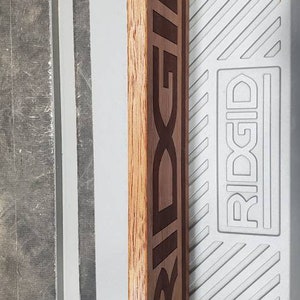 May include: A wooden sign with the word "RIDGID" engraved in black lettering. The sign is a dark brown color and has a natural wood grain.