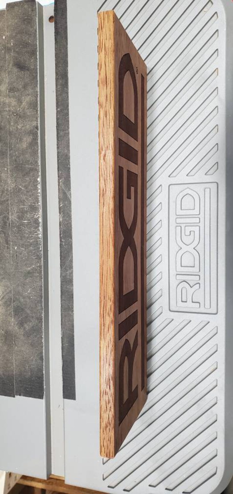 May include: A wooden sign with the word "RIDGID" engraved in black lettering. The sign is a dark brown color and has a natural wood grain.