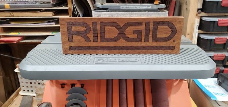 May include: A wooden sign with the word "RIDGID" engraved in a darker wood. The sign is sitting on a gray surface.