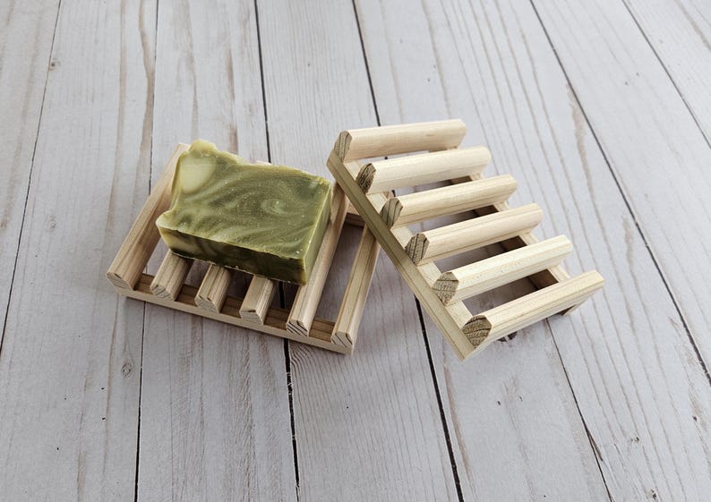 May include: A wooden soap dish with a bar of green and white soap on top. The soap dish is made of natural wood and has a slatted design.
