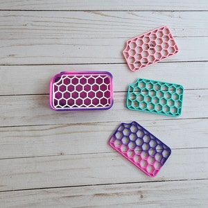 May include: Four silicone soap dishes in different colors and patterns. The dishes are shaped like honeycombs and are designed to drain water. The colors are pink, teal, purple, and white.
