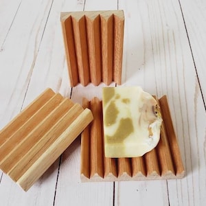 May include: Three wooden soap dishes with a natural finish. One dish holds a bar of soap with green and white tones. The dishes have a slatted design to allow for drainage. The soap dish is a bathroom accessory.